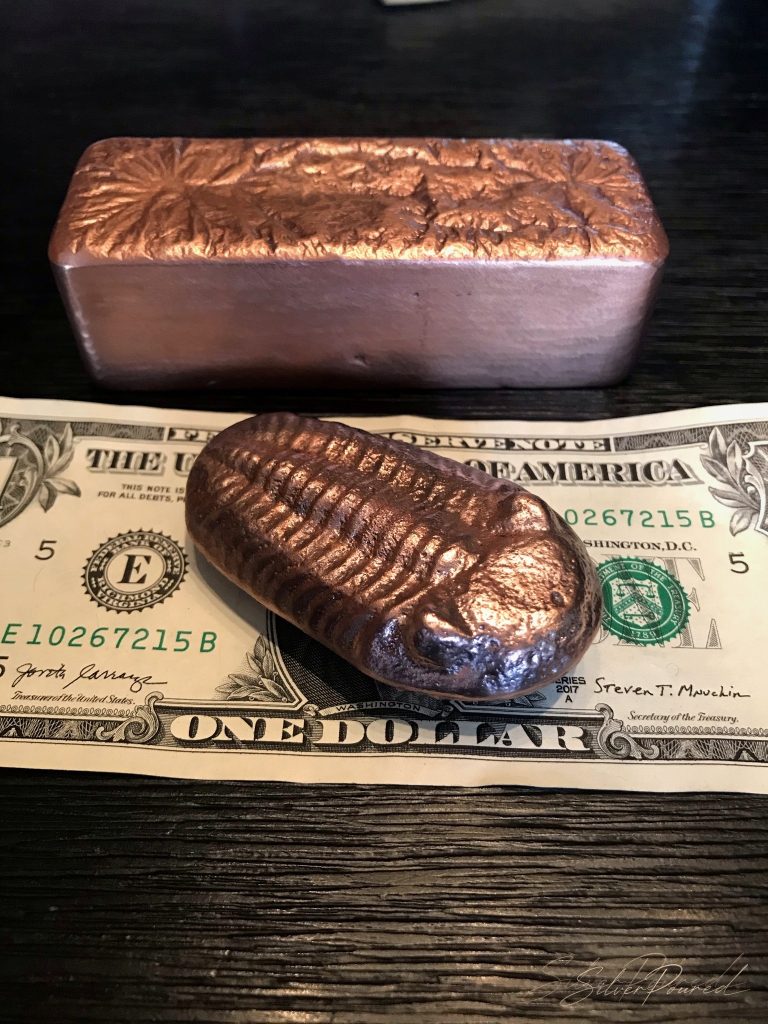 Hand-poured .999 copper bullion bar and trilobite copper pour displayed on a US dollar bill for scale on the SilverPoured Shop Copper page